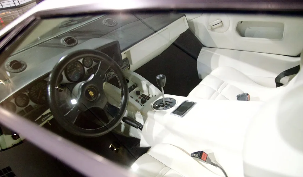 Car pedals visible in a garage footwell while a mechanic and driver discuss pedal feel