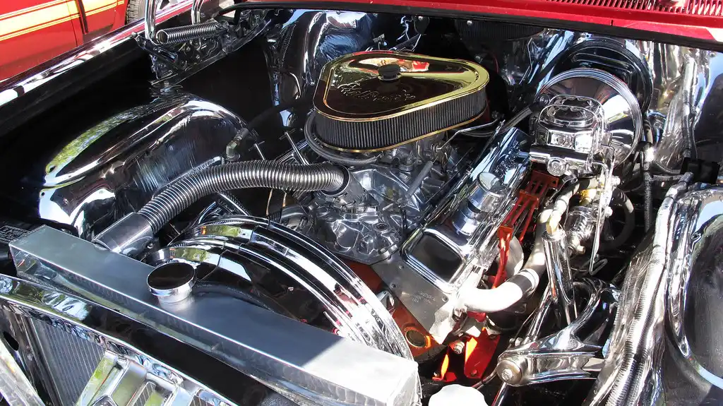 Older car owner comparing a carburetor and fuel injector setup beside a classic car