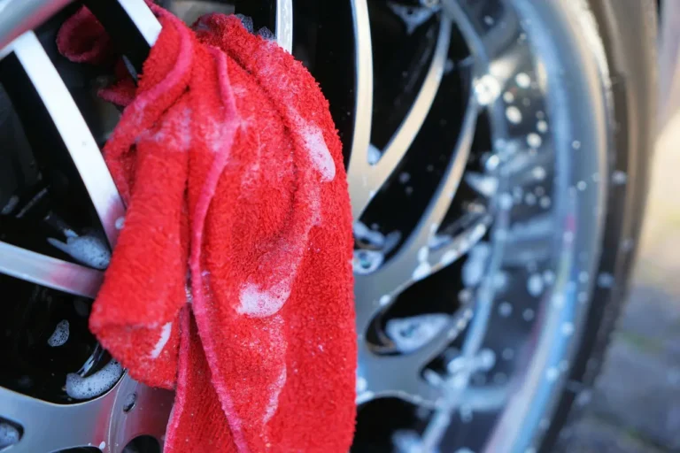 Person cleaning a car tire sidewall with a brush and soapy water beside a parked car