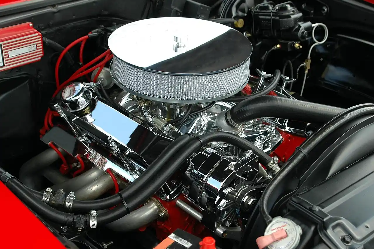 Person in a driveway checks a car maintenance list beside an open hood, tools, and fluid bottles