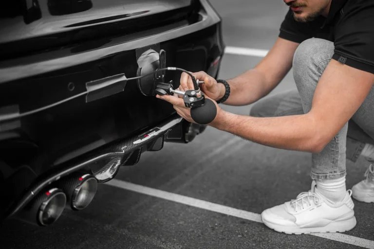 Mechanic inspecting a car's underside on a driveway while listening for a rattling noise.