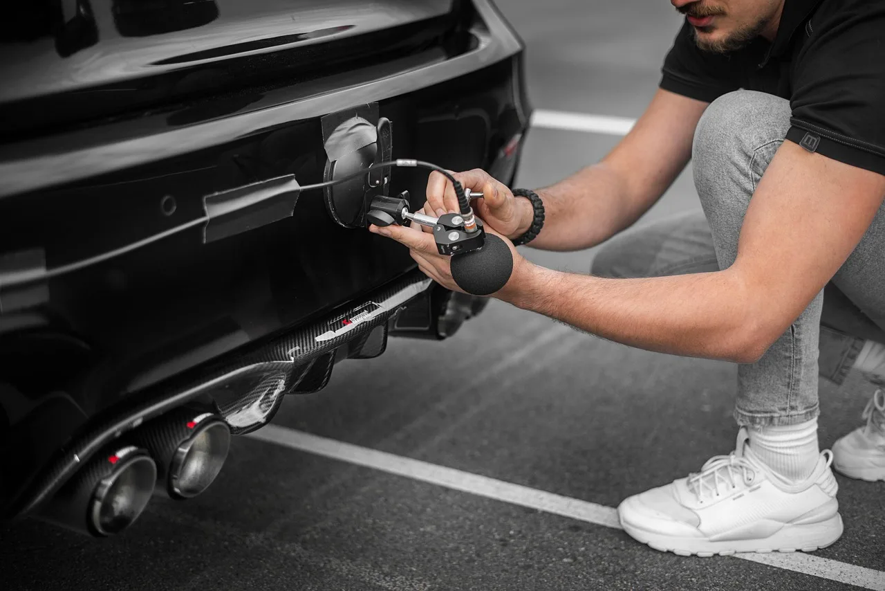 Mechanic inspecting a car's underside on a driveway while listening for a rattling noise.